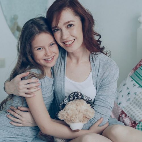 Portrait of adorable smiling girl embracing her happy mother and looking at camera together while sitting on bed in bright cozy bedroom at home