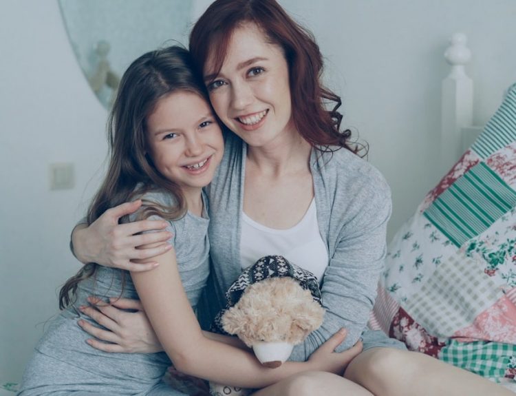 Portrait of adorable smiling girl embracing her happy mother and looking at camera together while sitting on bed in bright cozy bedroom at home