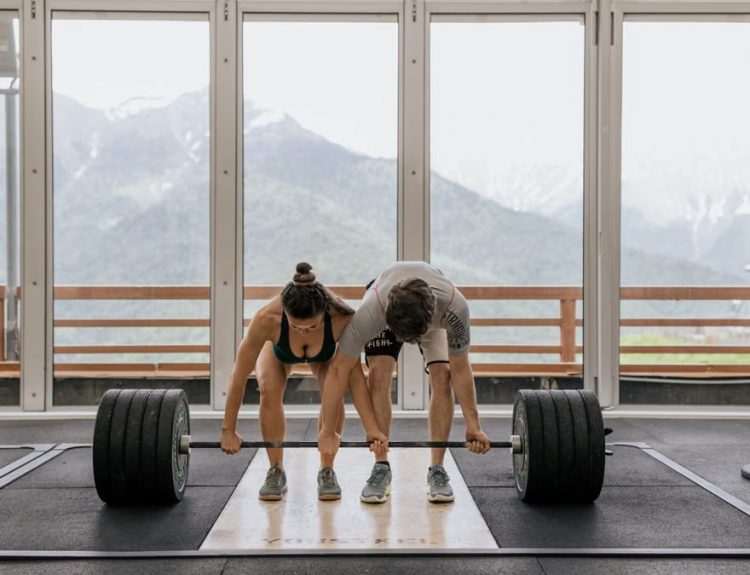 A couple lifting a heavy barbell together in a gym with mountain views.