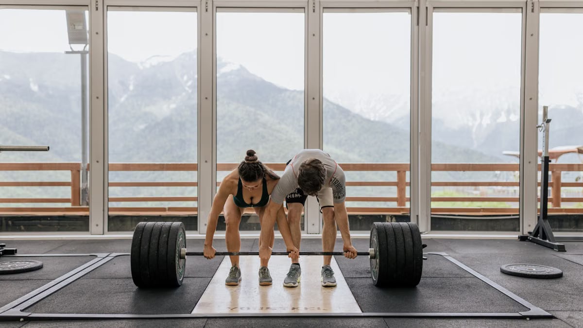 A couple lifting a heavy barbell together in a gym with mountain views.